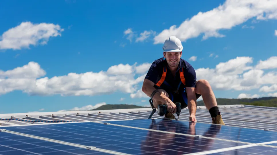 Monter pričvršćuje solarne kolektore na krov. Gleda u kameru i smije se.