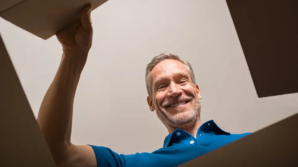 Sredovječni muškarac u plavoj polo majici, snimljen iz niskog kuta kao da je kamera u kutiji, s osmijehom gleda prema dolje dok otvara kartonsku kutiju na svijetloj pozadini.