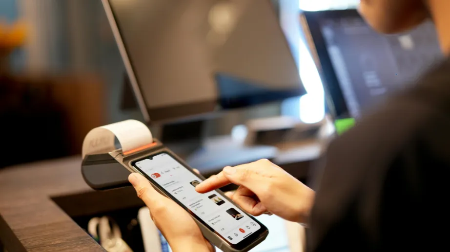 Osoba na blagajni koristi mobilni telefon za plaćanje, uz blagajnički terminal i računalni ekran, što prikazuje moderno beskontaktno plaćanje.