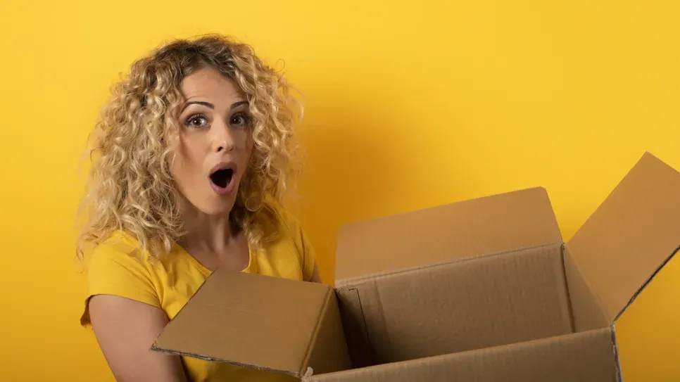 Kovrčava mlada žena u žutoj majici, s iznenađenim izrazom lica, gleda u otvorenu kartonsku kutiju na jednobojnoj žutoj pozadini.