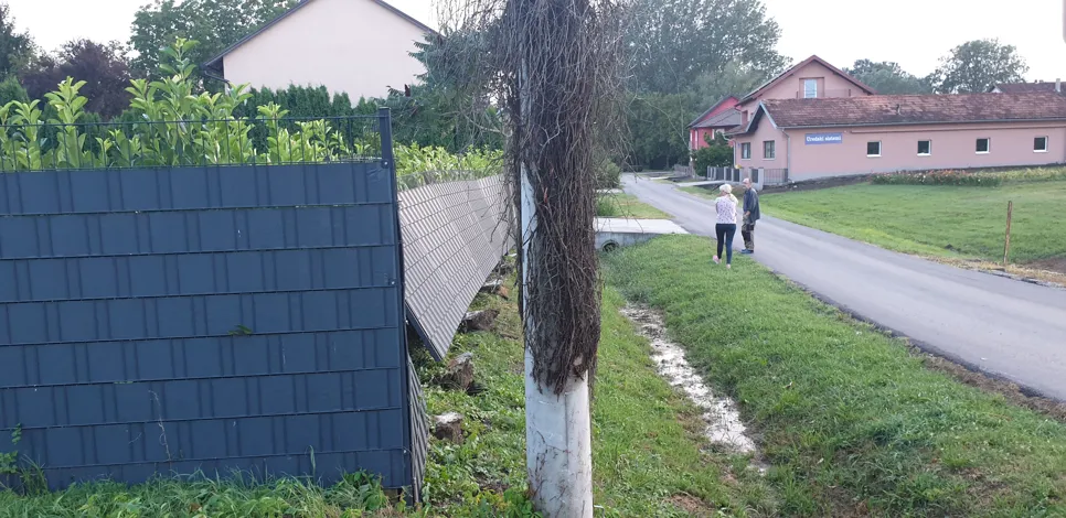 Leaning garden fence beside a rural road with houses and trees in the background, indicating structural instability.