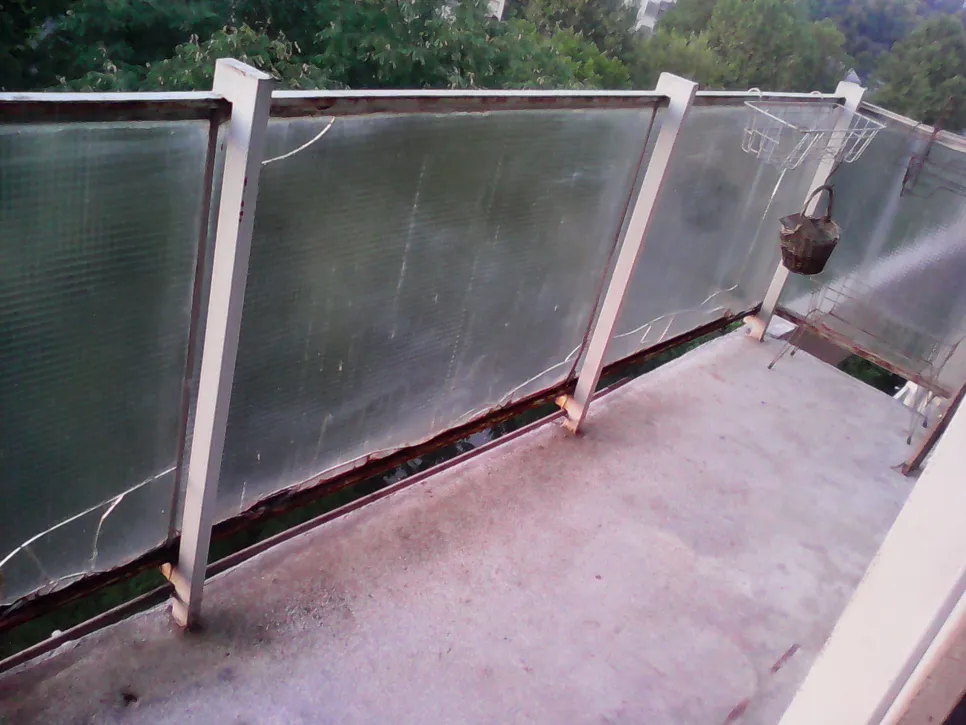 Balkon s puknutim i zahrđalim ogradnim panelima, koji prikazuju potencijalne sigurnosne opasnosti i potrebu za zamjenom.