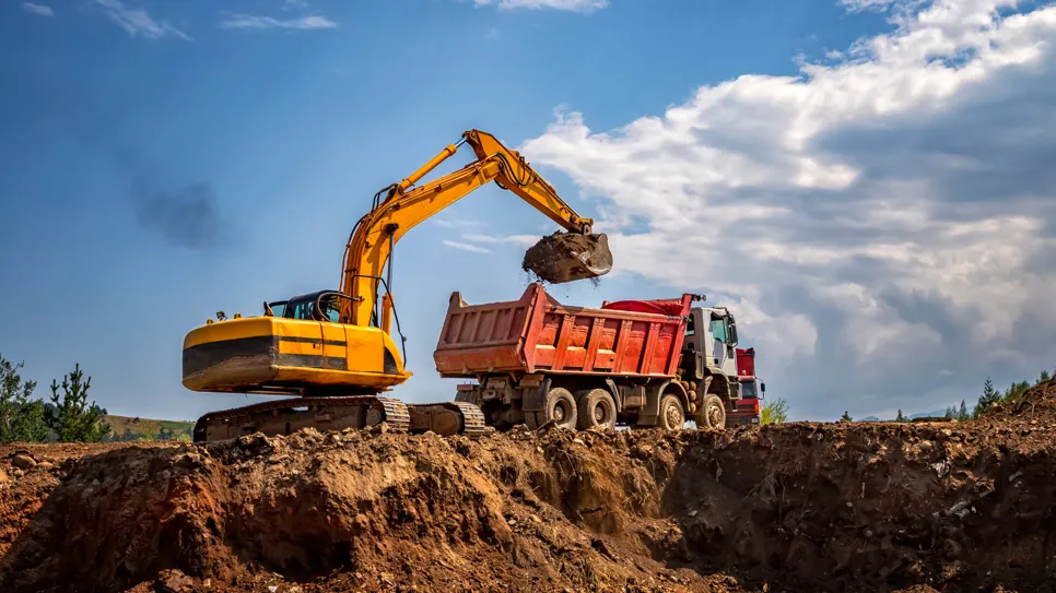 Žuti bager prenosi zemlju u crveni kamion na gradilištu.