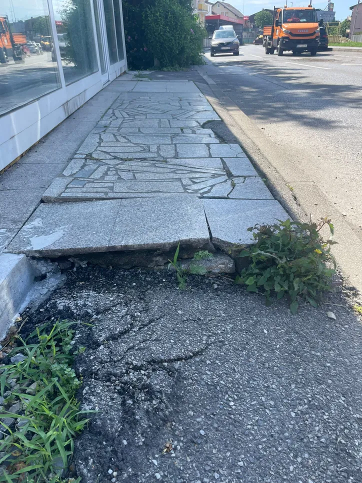 Cracked sidewalk with damaged surface and growing weeds alongside a road, indicating pavement issues.