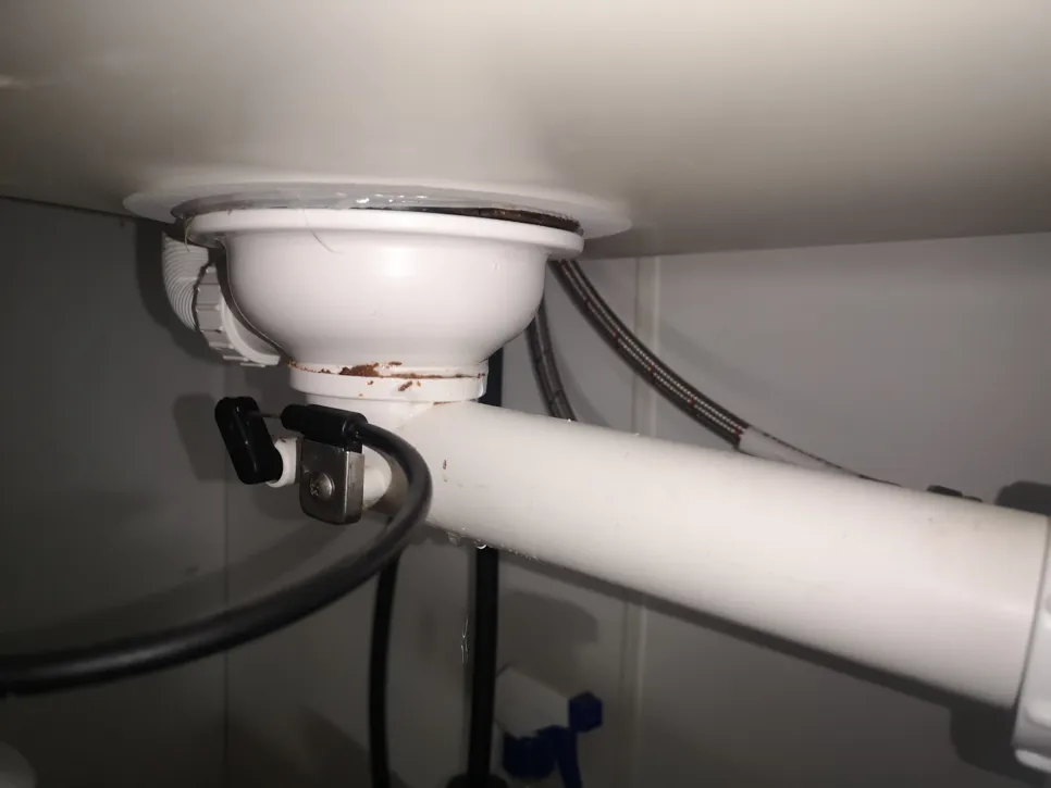 Kratak prikaz cijevi ispod kuhinjskog sudopera koja curi, s vidljivom hrđom i kapljicama vode ispod, ističući probleme s vodovodom.