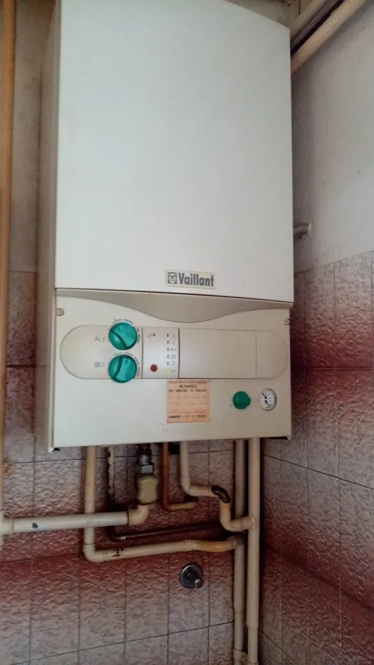Plinski bojler Vaillant s složenim sustavom cijevi u kutu prostorije s pločicama, s kontrolnim gumbima i manometrom.