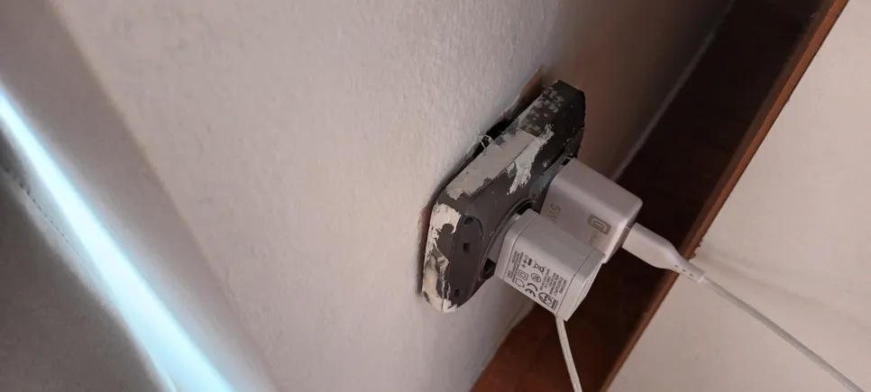 Zidna utičnica s vidljivim oštećenjem i izloženim rubovima, s priključenim USB punjačem.