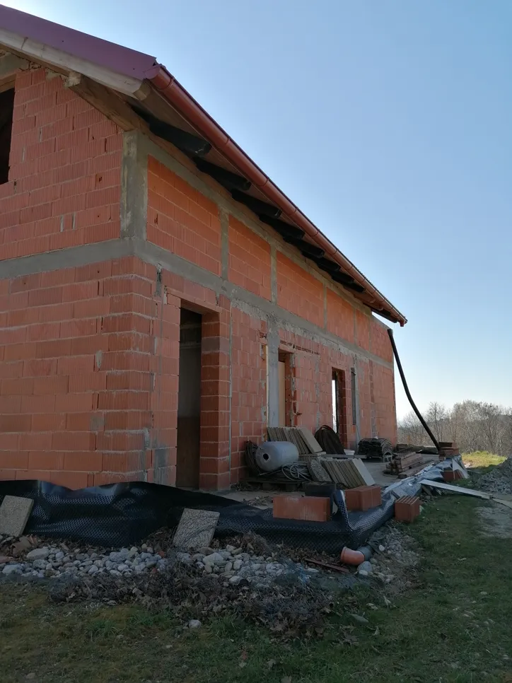 Nedovršena kuća od opeke s nepotpunim krovom i oblogom, prikazuje otvorene prostore i građevinski materijal u blizini.