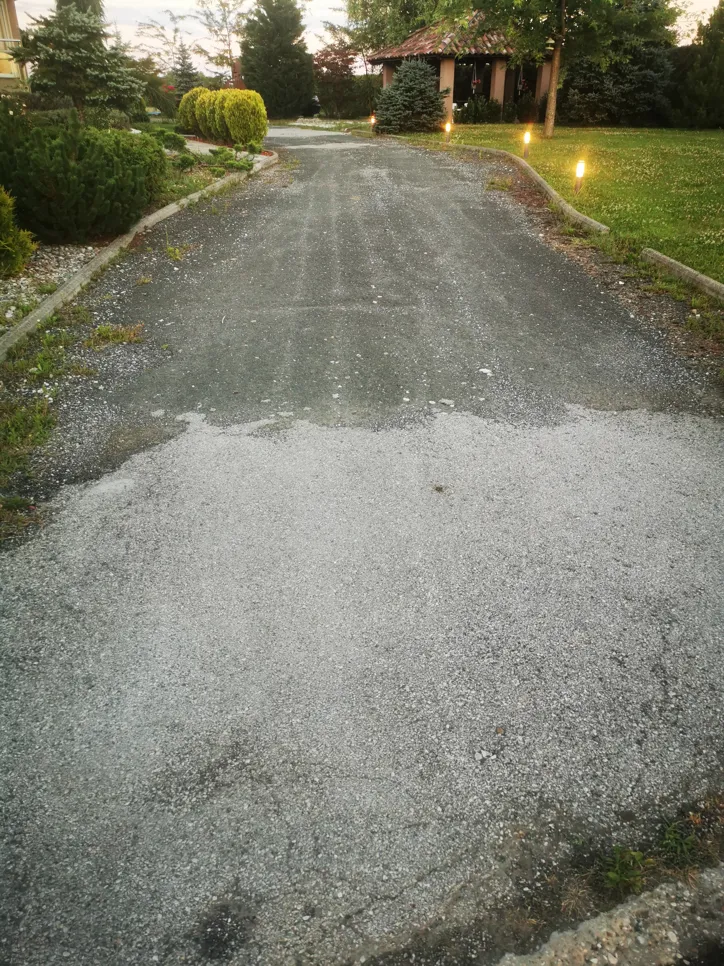 Gravel driveway showing signs of wear and uneven surface, surrounded by neatly trimmed bushes with pathway lights.