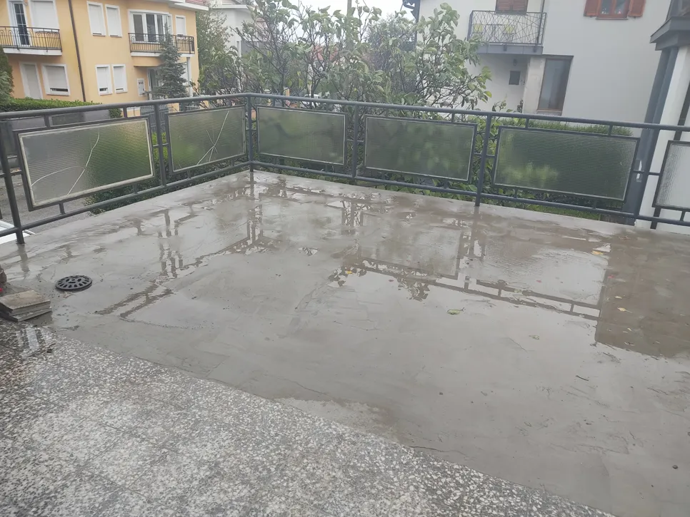 A balcony with standing water due to inadequate drainage after rainfall, surrounded by buildings and plants.