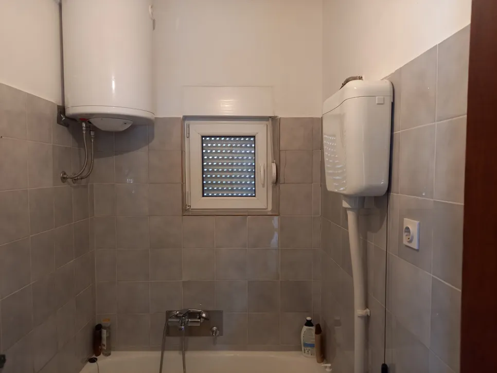 Kupaonica s zidnim bojlerom i pločicama iznad kade, s malim prozorom s roletama.