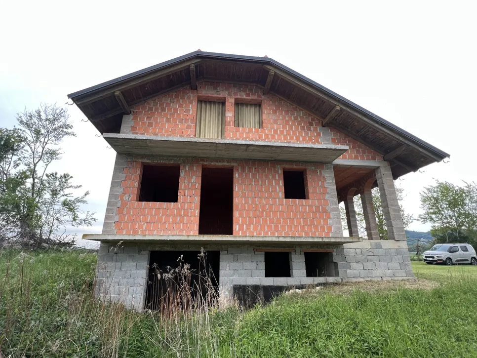 Nedovršena kuća od opeke s izloženom strukturom i otvorenim prozorima, smještena u travnatoj površini.