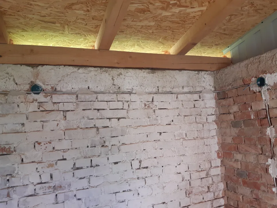 Izloženi električni kablovi na obojenom zidu od opeke u prostoriji s nedovršenim drvenim stropom.