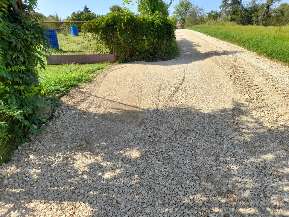 Gravel driveway with uneven surface and ruts, surrounded by greenery, under clear sky with visible tire tracks.