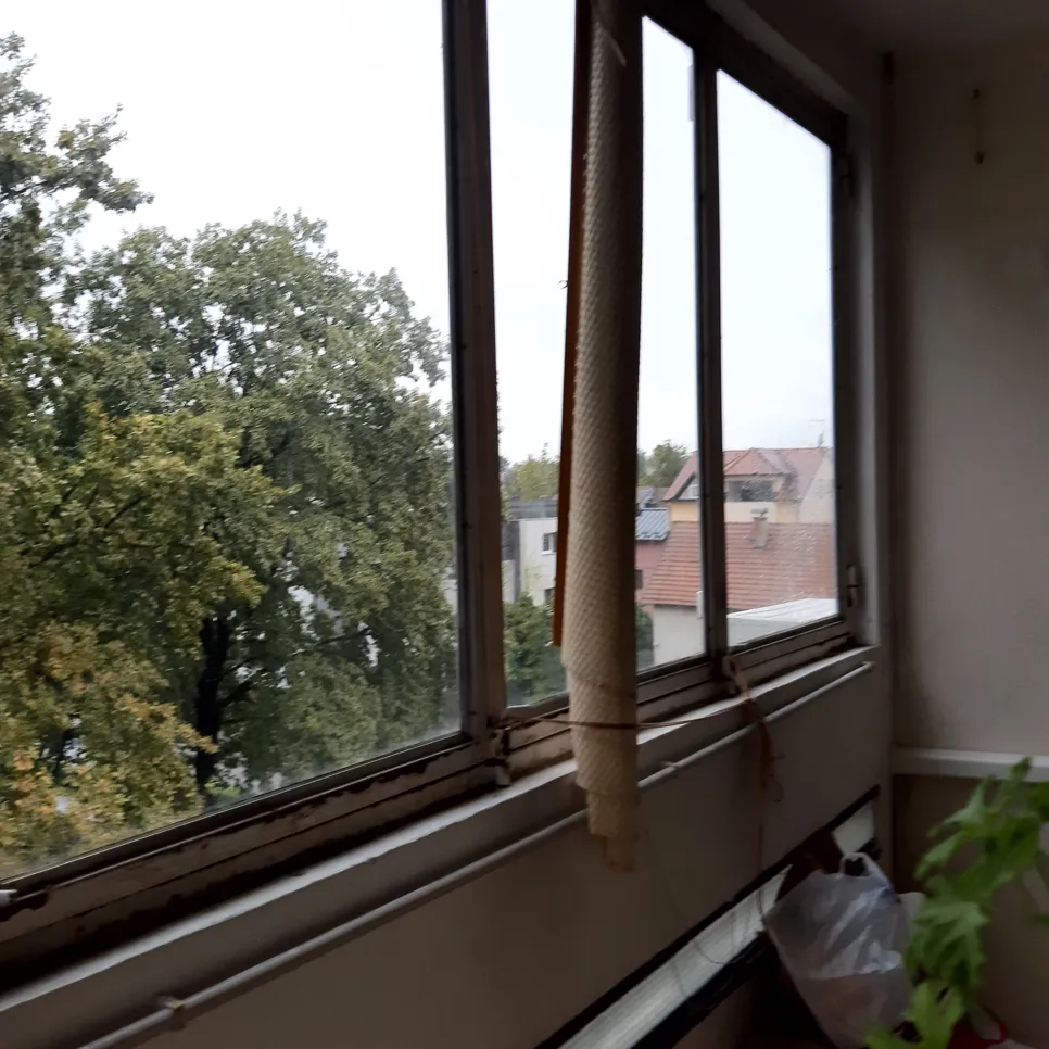 Hrđavi okvir stambenog prozora s djelomično otvorenom zavjesom koja gleda na drveće i krovove.