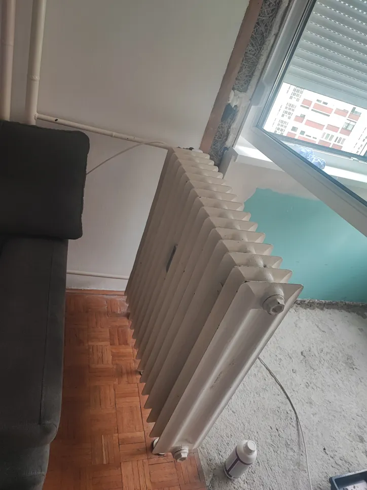 Stari radijator postavljen pored otvorenog prozora s nedovršenim zidovima koji pokazuju moguću neučinkovitost grijanja.