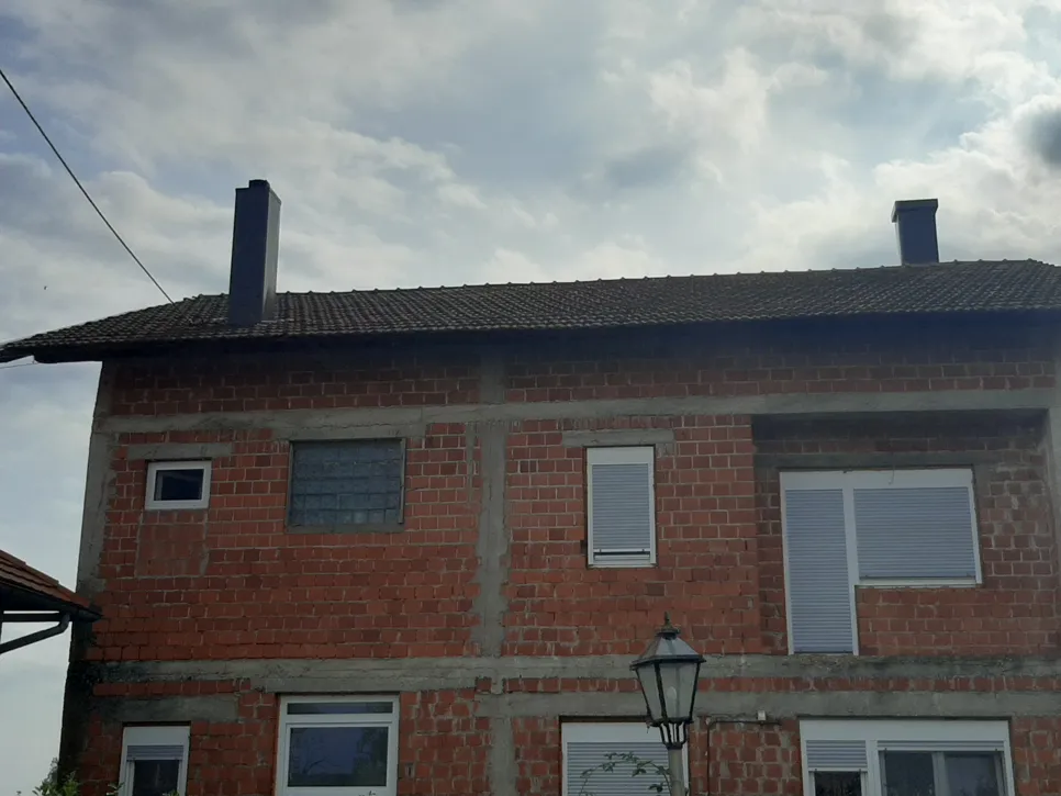 Nedovršena kuća od opeke prikazuje izložene zidove i dovršen krov od šindre, dimnjak vidljiv protiv oblačnog neba.