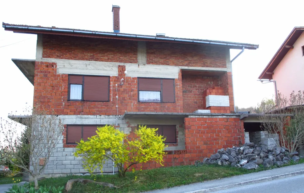 Kuća od opeke s djelomično nedovršenom vanjštinom, koja prikazuje izložene opeke i beton. Smještena u predgrađu.