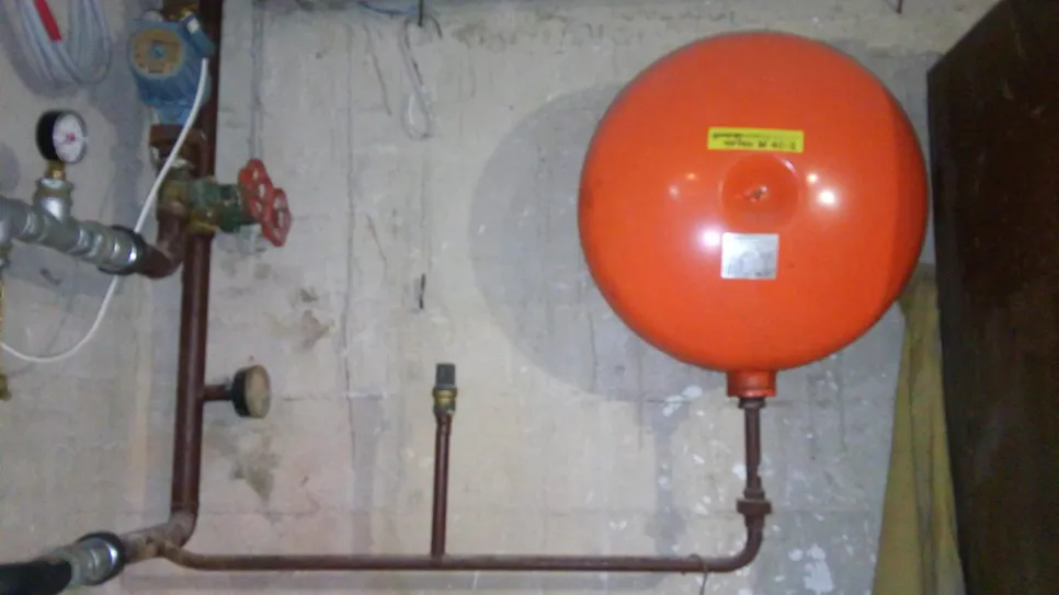 Narančasti ekspanzijski spremnik povezan s kućnim sustavom grijanja, prikazujući cijevi i ventile.