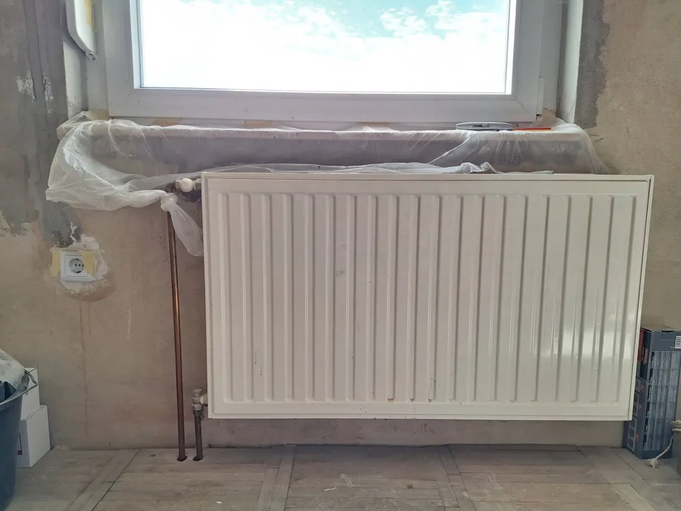 Radijator montiran na zid ispod prozora u prostoriji koja se renovira, s vidljivim bakrenim cijevima.