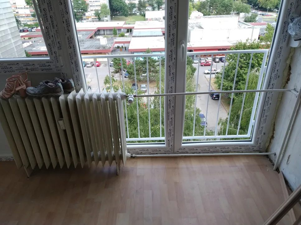 Radijator je postavljen ispod velikog prozora s vanjskom zaštitnom ogradom, s pogledom na zgrade i zelenilo.