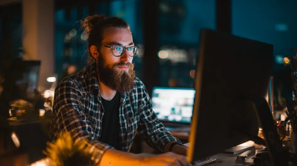 Koncentrirani bradati muškarac s naočalama radi na računalu kasno navečer, simbolizirajući osobno vrijeme i trud koji doprinose trošku posjedovanja web stranice. Njegova predanost ilustrira "uloženi trud" potreban za izgradnju i održavanje uspješne online prisutnosti.